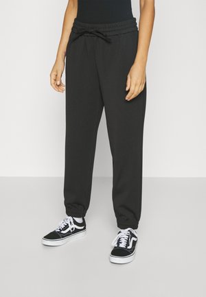 Person, die schwarze Jogginghosen mit Kordelzug und schwarz-weiße Sneaker trägt, steht auf einem hellgrauen Boden vor einer weißen Wand.