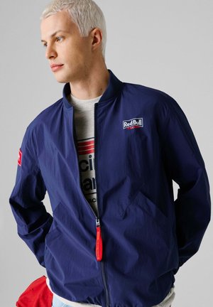 Jonge man met kort platinumblond haar, gekleed in een donkerblauw Red Bull Racing-jasje over een grijs grafisch T-shirt, kijkend naar links.