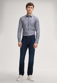 Langarmige button-up shirt in blauw met een bloemenpatroon, gecombineerd met donkere spijkerbroek en witte sneakers. Model staat voor een neutrale achtergrond.