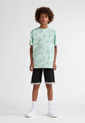 Tricou verde mentă cu mâneci scurte și imprimeuri botanice, asortat cu pantaloni scurți negri cu tiv răsucit și adidași albi.