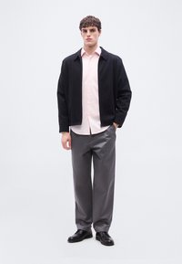 Νεαρός άνδρας που φοράει μαύρο μπουφάν, ανοιχτόροζ πουκάμισο με κουμπιά, γκρι παντελόνι φαρδιάς γραμμής και μαύρα επίσημα παπούτσια, στέκεται μπροστά σε λευκό φόντο.