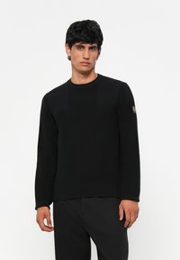 Pulover negru din material cu nervuri, cu mâneci lungi, decolteu rotund și detalii subtile cu panouri; asortat cu pantaloni negri. Prezintă un badge cu logo pe mânecă.