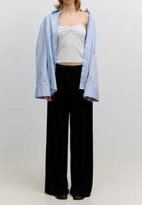 Lichtblauwe oversized shirt, witte aansluitende tanktop en losse zwarte broek met een trekkoord in de taille en een gladde stoftextuur.