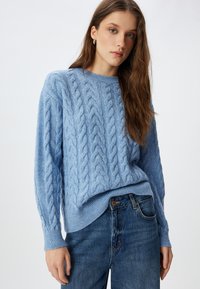 Maglione a coste di colore azzurro chiaro con scollatura rotonda e polsini a coste, abbinato a jeans blu scuro. Texture morbida e vestibilità rilassata.