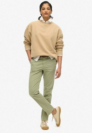 Sweatshirt beige à coupe ample sur une chemise à col blanc, associé à un pantalon vert clair et des baskets beiges avec des semelles texturées.