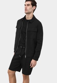 Male model wearing a black zip-up jacket with chest pockets, black T-shirt, and black drawstring shorts, standing against a plain background.