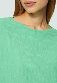 Top verde menta realizzato in tessuto testurizzato con dettagli arricciati, caratterizzato da un collo rotondo e maniche corte a raglan.