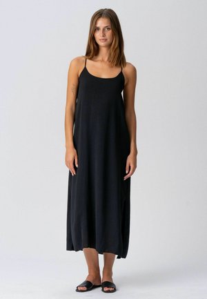 Vállig érő barna hajú nő laza, fekete ujjatlan maxi ruhában és fekete papucsban, sima háttér előtt áll.