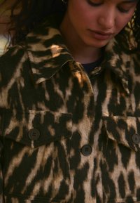 Harmaa eläinkuosilla varustettu takki vaaleanruskeassa ja tummanvihreässä värissä. Takissa on kauluskäärme, kaksi rintataskua ja tummista nappeista valmistettu kiinnitys.