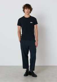 Čierne bavlnené tričko s malým bielym logom na hrudi, kombinované s voľnými tmavými nohavicami a čiernymi šnurovacími topánkami. Minimalistický dizajn.