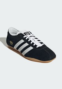 Melns zamšādas sporta apavi ar trim baltiem svītrojumiem, auklu dizains, gumijas zole un zelta "adidas Tokyo" logo sānos.