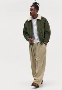 Zielona kurtka typu bomber z sztruksu z białym sztucznym kołnierzem z kożucha, połączona z szerokimi beżowymi spodniami ze sztruksu i brązowymi butami wsuwanymi.