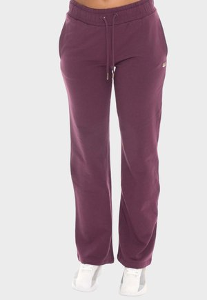 Vrouw draagt paarse joggingbroek met trekkoord en witte sneakers, staand tegen een neutrale achtergrond.