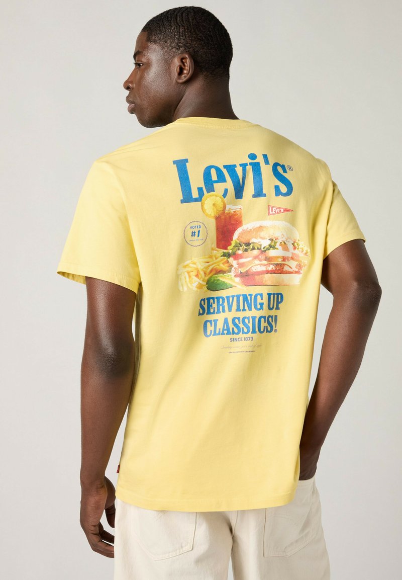 Mann trägt ein gelbes Levi's T-Shirt mit blauem Text und einer Grafik von Burger, Pommes und Getränk, schaut nach links vor neutralem Hintergrund.
