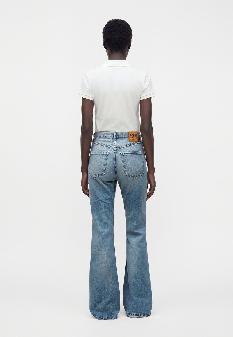 Personne debout de dos portant un polo blanc à manches courtes et un jean bleu clair évasé taille haute avec des poches à l'arrière.