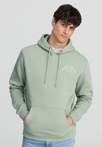 Hoodie verde menta con tasca a marsupio frontale, cappuccio con cordino e stampa grafica "North Carolina". Materiale morbido con polsini a costine.