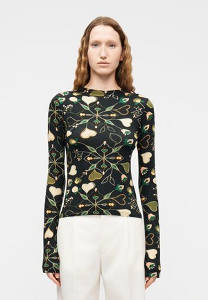 Jonge vrouw met lang golvend haar die een nauwsluitende zwarte lange mouw top draagt, bedrukt met groene en beige hart- en bloemmotieven, gecombineerd met witte broek.