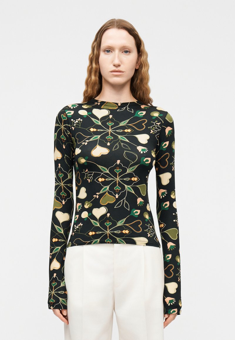 Νεαρή γυναίκα με μακριά κυματιστά μαλλιά που φοράει εφαρμοστή μαύρη μπλούζα με μακριά μανίκια, διακοσμημένη με πράσινα και μπεζ μοτίβα καρδιών και λουλουδιών, σε συνδυασμό με λευκό παντελόνι.