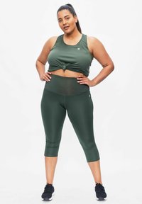 Olivgrünes Sport-Crop-Top mit Knoten-Detail an der Vorderseite, kombiniert mit hochgeschnittenen Leggings. Glattes, elastisches Material; nahtloses Design.