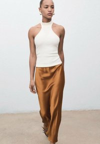 Bílý halter top v kombinaci s dlouhou, splývavou sukní z rustikálového saténu. Modelka má na sobě sandály na podpatku. Pozadí je jednoduchá světlá zeď.