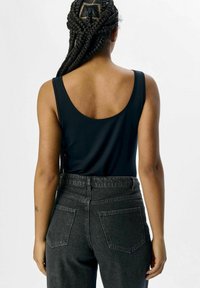 Schwarzes ärmelloses Top mit abgerundetem Rücken, kombiniert mit hoch taillierten dunkelgrauen Jeans. Glatte Stoffstruktur mit normalen Taschen.