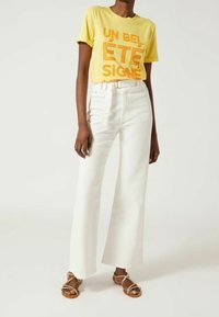 Žluté tričko s krátkým rukávem a výrazným oranžovým textem; bílé vysoké rozšířené džíny; hnědé sandály na plochém podpatku s pásky.