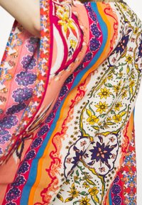 Tissu coloré avec des motifs floraux et géométriques dans des tons d'orange, de rose, de violet, de jaune, de bleu et de blanc, maintenu par le bras d'une personne.