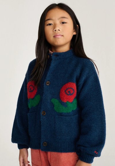 Jeune fille aux longs cheveux foncés porte un cardigan bleu boutonné avec des motifs floraux rouges et verts et un pantalon orange.