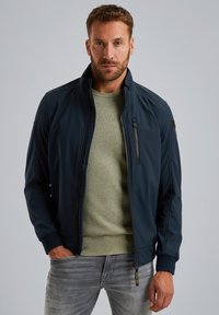 Marineblaue Bomberjacke mit hohem Kragen, Front-Reißverschlusstasche und gerippten Bündchen, kombiniert mit einem hellgrünen strukturierten Pullover und grauen Jeans.