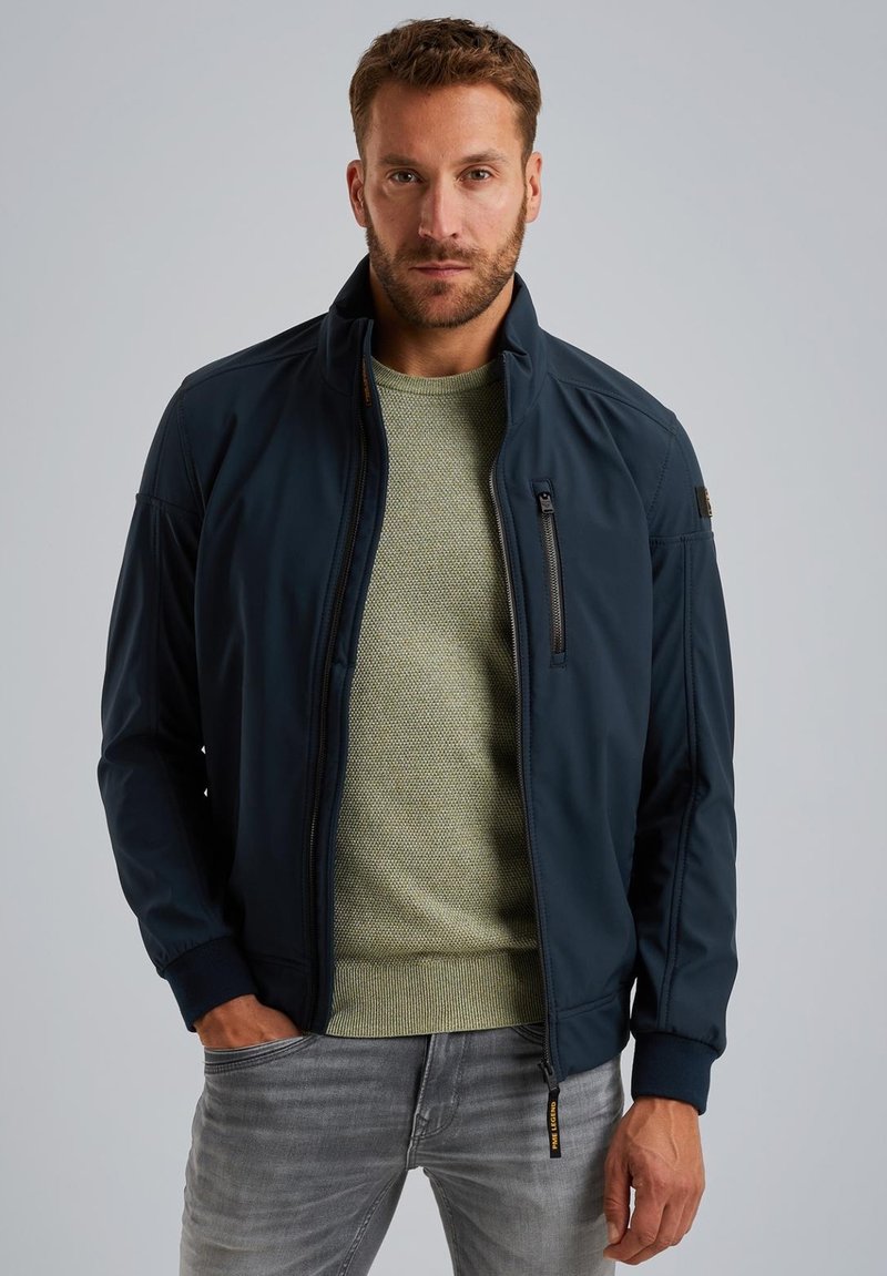 Marineblaue Bomberjacke mit hohem Kragen, Front-Reißverschlusstasche und gerippten Bündchen, kombiniert mit einem hellgrünen strukturierten Pullover und grauen Jeans.