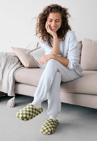 Cozy, pale blue ribbed loungewear with long sleeves and pants. Plush green and white checkered slippers. Sofas and textured blankets in background.