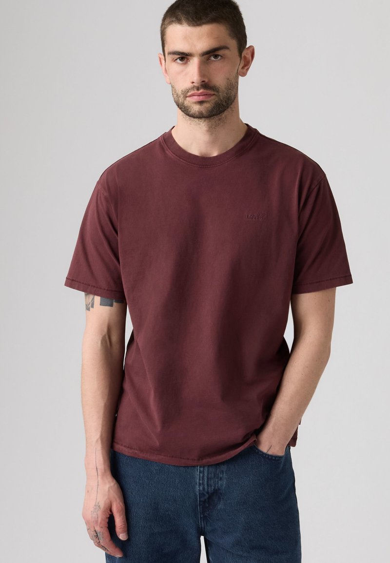 Uomo con capelli corti e scuri e barba che indossa una maglietta bordeaux semplice e jeans blu, in piedi con una mano in tasca.