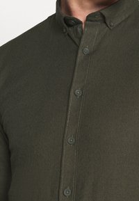 Camicia verde scuro con bottoni, caratterizzata da un colletto strutturato, tessuto testurizzato e cuciture a contrasto. I bottoni sono scuri con una finitura liscia.