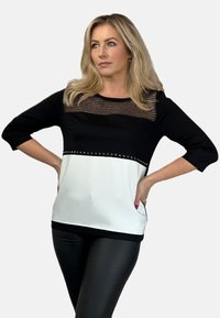 Czarny i biały top z siateczkowym panelem i dekoracyjnymi ćwiekami wzdłuż dolnego szwu. Posiada rękawy o długości trzy czwarte i gładkie wykończenie.