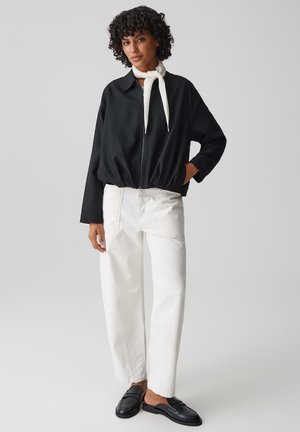 Donna con capelli ricci indossa una giacca nera con cerniera, una sciarpa bianca, pantaloni bianchi a gamba larga e mocassini neri slip-on su sfondo grigio.