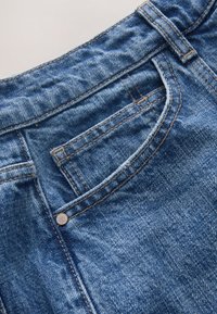 Nahaufnahme einer Brusttasche aus blauem Denim mit sichtbaren Nähten, Gürtelschlaufe und Metallniete auf texturiertem Stoff.