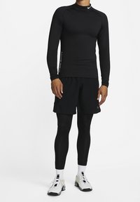 Mann trägt ein schwarzes langärmeliges Kompressionsshirt, schwarze Shorts über schwarzen Leggings, weiße Socken und weiße Sportschuhe auf weißem Hintergrund.