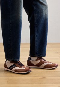 Sneakers in pelle marrone e rete con accenti scuri, pannelli testurizzati e un logo. Indossati con pantaloni in denim scuro su un pavimento di legno.