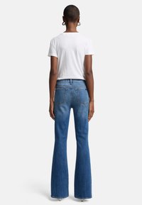 Jean flare bleu avec un délavage moyen, à taille haute, avec cinq poches, associé à un T-shirt blanc, vu de dos.