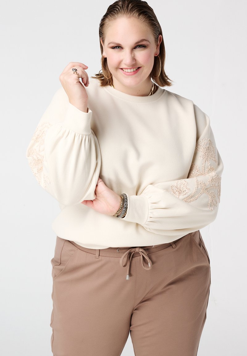 Roomkleurige sweatshirt met volumineuze mouwen en beige borduursel. Gecombineerd met bruine broek en gelaagde armbanden. Soepele textuur.