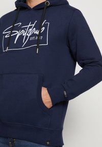 Marineblauwe hoodie met een voorzak, trekkoordsluiting en een witte logo-opdruk. Ribgebreide mouwen en zoom, zacht materiaal.