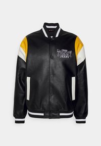 Musta nahkainen varsity-takki, jossa on keltaisia, valkoisia ja mustia yksityiskohtia. Takissa on resorit hihansuissa sekä rintataskussa logo, jossa on nappikiinnitys.