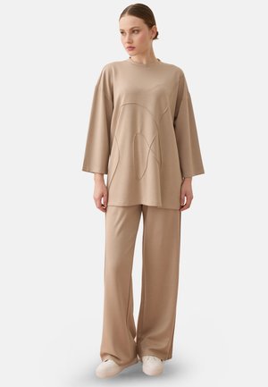 Kvinde iført løs beige langærmet top med abstrakt syning og matchende vidde bukser, stående og kiggende til siden.