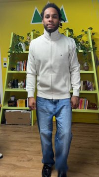 Felpa grigia chiaro con cerniera e collo alto, abbinata a jeans di denim blu e scarpe nere, su uno sfondo giallo con scaffali verdi.