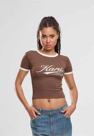 Joven mujer con el pelo trenzado que lleva un crop top marrón con ribete blanco y texto "Kani", combinado con vaqueros azules, con las manos en los bolsillos.