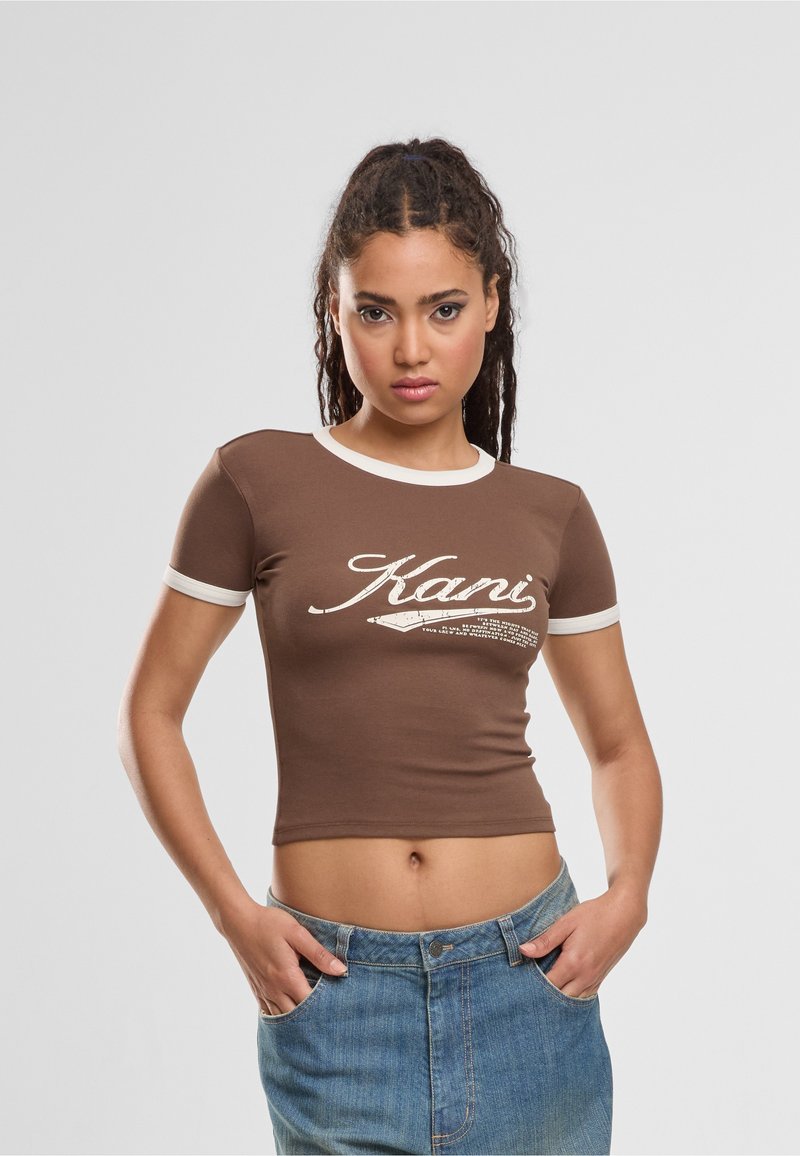 Νεαρή γυναίκα με πλεξούδες που φοράει καφέ crop top με λευκή επένδυση και το κείμενο "Kani", συνδυασμένο με μπλε τζιν, με τα χέρια στις τσέπες.