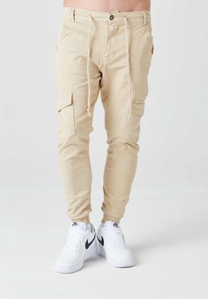 Beige cargobroek met trekkoord in de taille, gedragen met witte Nike Air Force 1 sneakers met zwarte swoosh-logo's.