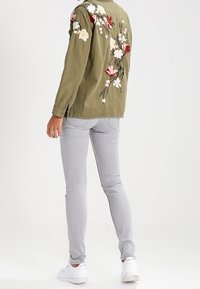 Olivově zelená bunda s květinovou výšivkou na zádech, spárovaná s světlými šedými skinny džíny a bílými teniskami. Hladké textury a přiléhavý design.