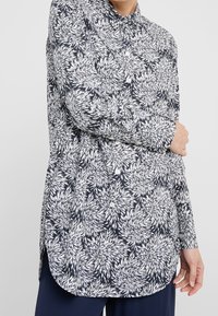 Camicia a maniche lunghe di colore blu navy con un motivo floreale bianco, dotata di colletto a punta e orlo curvo. Realizzata in tessuto leggero.