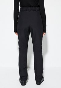 Zwarte broek met een hoge elastische tailleband, taps toelopende pijpen en een zachte textuur. Gecombineerd met zwarte schoenen die zichtbaar zijn aan de onderkant.